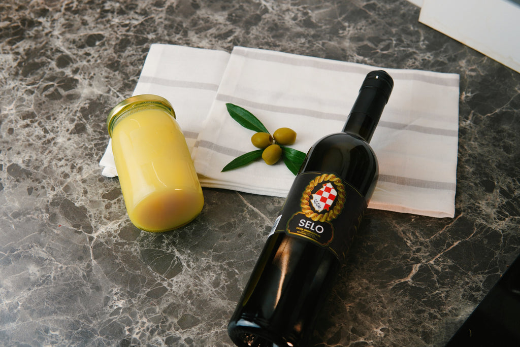 A black bottle of Croatian olive oil and a jar of creamy beef tallow side by side on a marable countertop, accented with fresh olive sprigs.