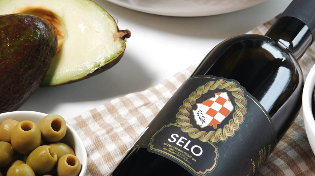 A bottle of Selo Olive oil positioned in the foreground, accompanied by a captivating assortment of marinated olives and sliced avocadoes in the background, creating a captivating tableau of Mediterranean flavors and healthy indulgence.