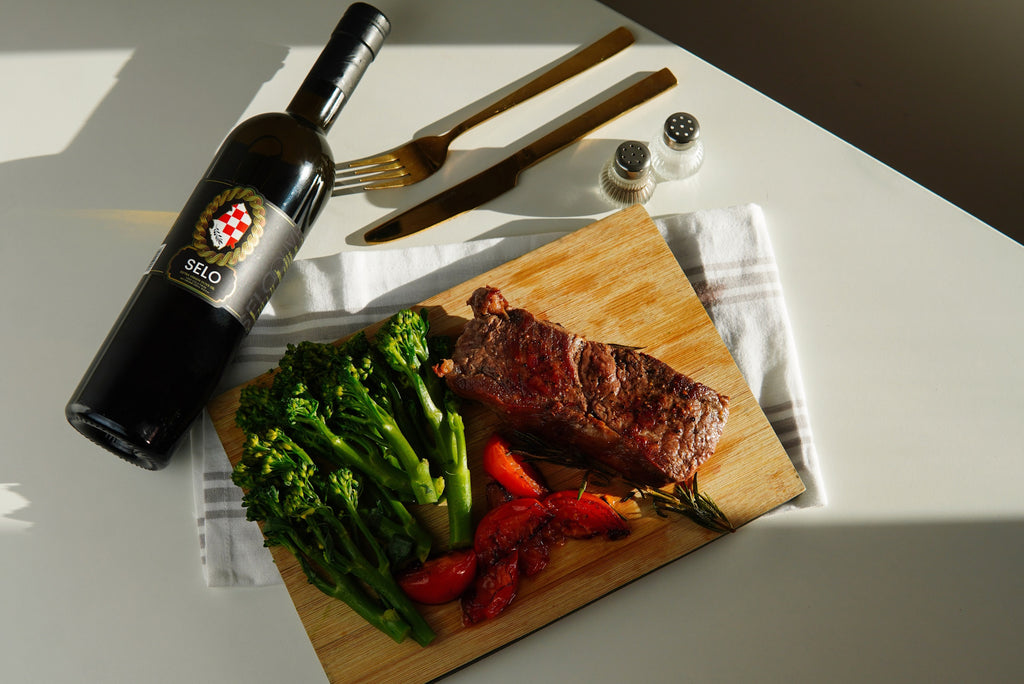 Perfectly cooked steak resting on a wooden cutting board, garnished with rosemary, with a bottle of Selo Croatian Olive Oil beside it.